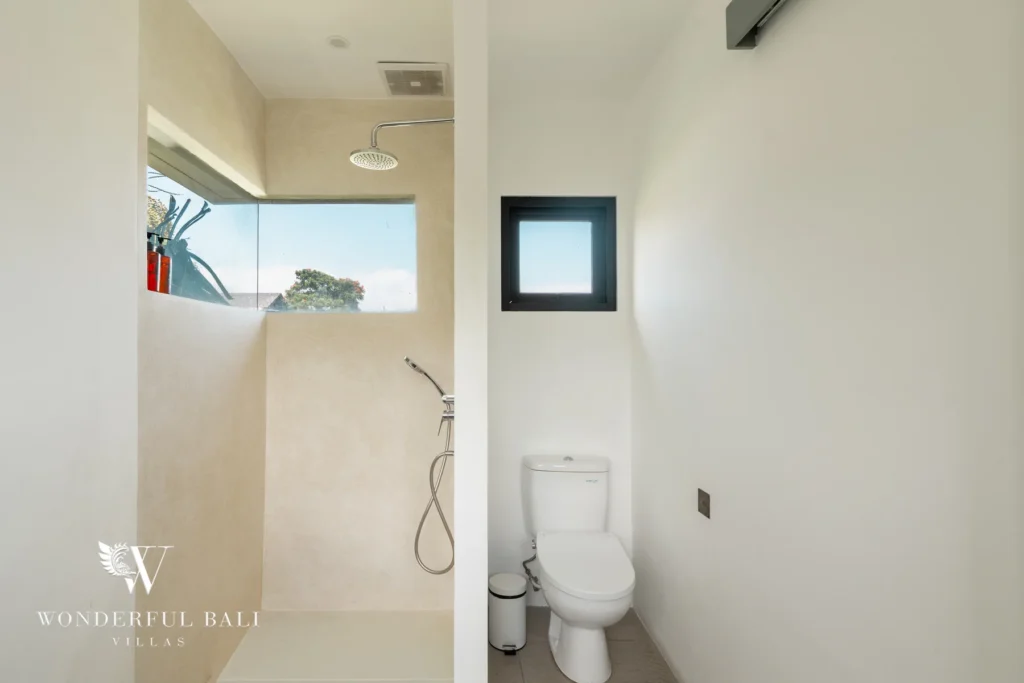 A bathroom with a walk-in rain shower, handheld sprayer, toilet and a small window looking onto greenery at a 3-bedroom villa in Berawa, Bali