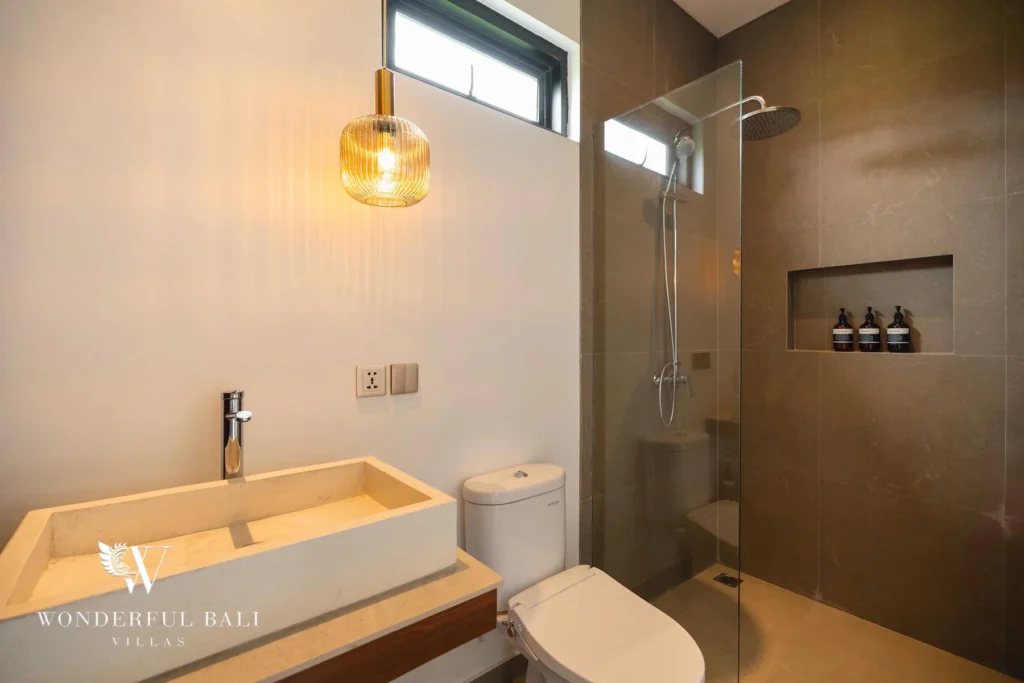 An en-suite bathroom with a stone vessel sink, a glass walk-in shower with grey tiling and a hanging amber pendant light at a 4-bedroom villa in Canggu, Bali
