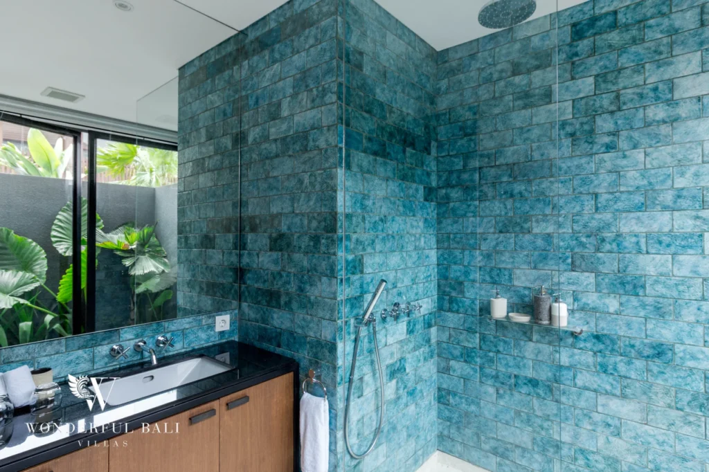 A bathroom with teal brick-tiled walls, a rain shower and a timber vanity beside a window onto tropical foliage at a 4-bedroom villa in Batu Bolong, Canggu, Bali