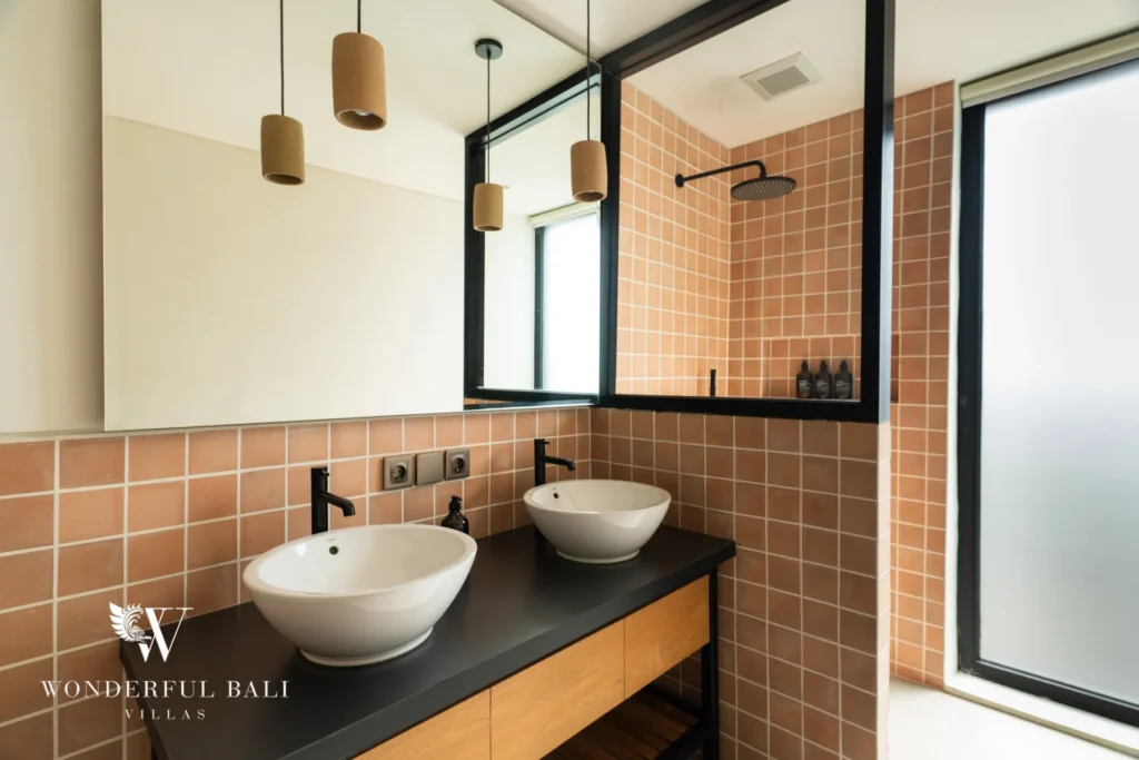An en-suite bathroom clad in terracotta tiles with twin vessel sinks on a black vanity, a walk-in rain shower and clay pendant lights at a 5-bedroom villa in Batu Bolong, Canggu, Bali