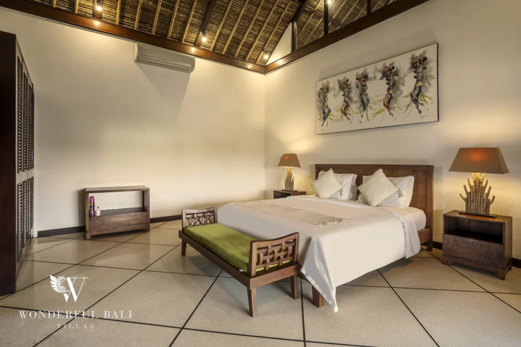 A king bedroom with a high thatched vaulted ceiling, wooden bench and framed artwork above the bed at a 4-bedroom villa in Seminyak, Bali
