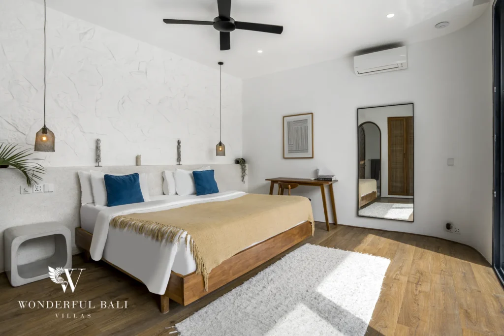Bedroom with a wooden bed, blue cushions, a textured white headboard wall, a desk and a full-length standing mirror at a 3-bedroom villa in Bingin, Uluwatu, Bali