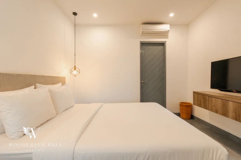 A bedroom with a white double bed, geometric pendant light, wall-mounted smart TV on a wooden console and air conditioning at a 3-bedroom villa in Berawa, Bali
