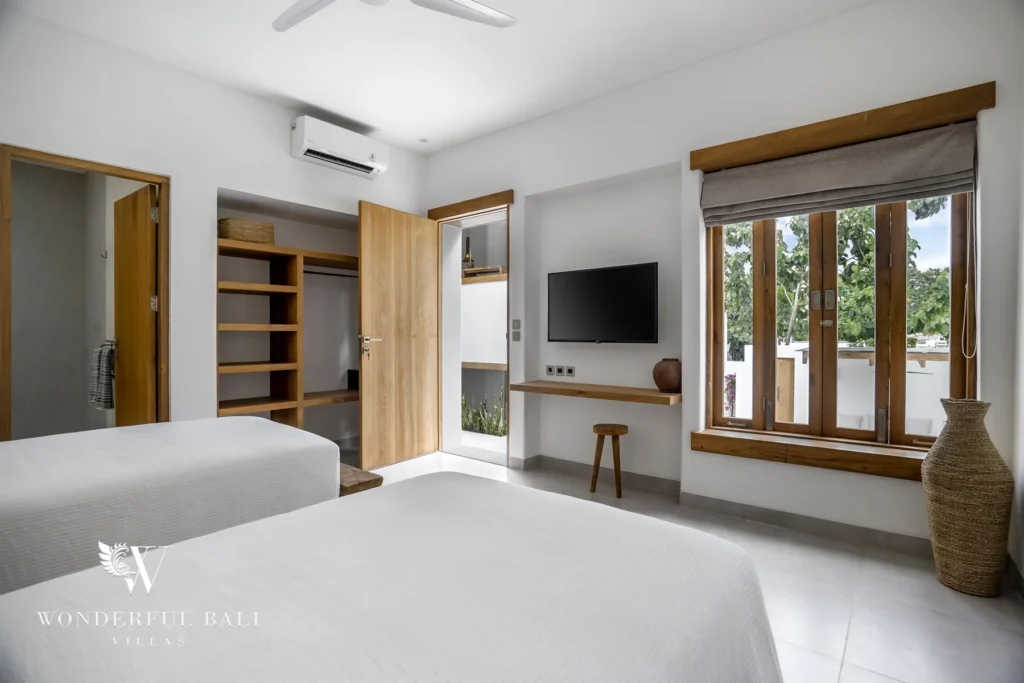 Twin beds face a wall-mounted TV, a small wooden desk stool and open wardrobe shelving beside a bright window and a door to the courtyard at a 3-bedroom villa in Bingin, Uluwatu, Bali