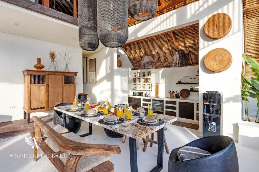 A marble dining table set for a meal sits beneath dark woven pendant lamps beside an open kitchen and antique wood cabinet under a high ceiling at a 4-bedroom villa in Canggu, Bali