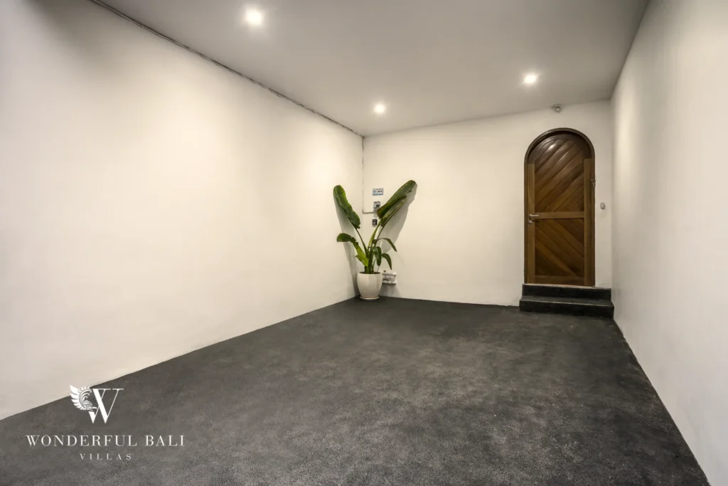 A minimalist entry space with grey carpet, white walls, a potted plant and an arched wooden door at a 3-bedroom villa in Bingin, Uluwatu, Bali
