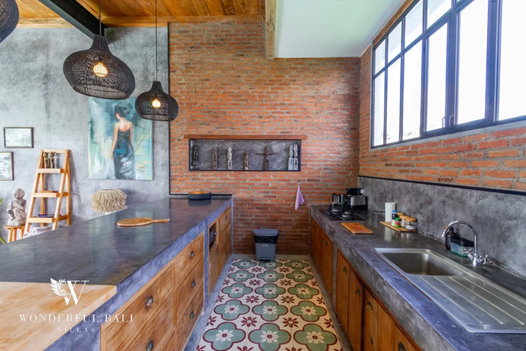 A galley kitchen with exposed brick wall, concrete countertops, timber cabinets, patterned floor tiles and woven pendant lights at a 3-bedroom villa in Seminyak, Bali