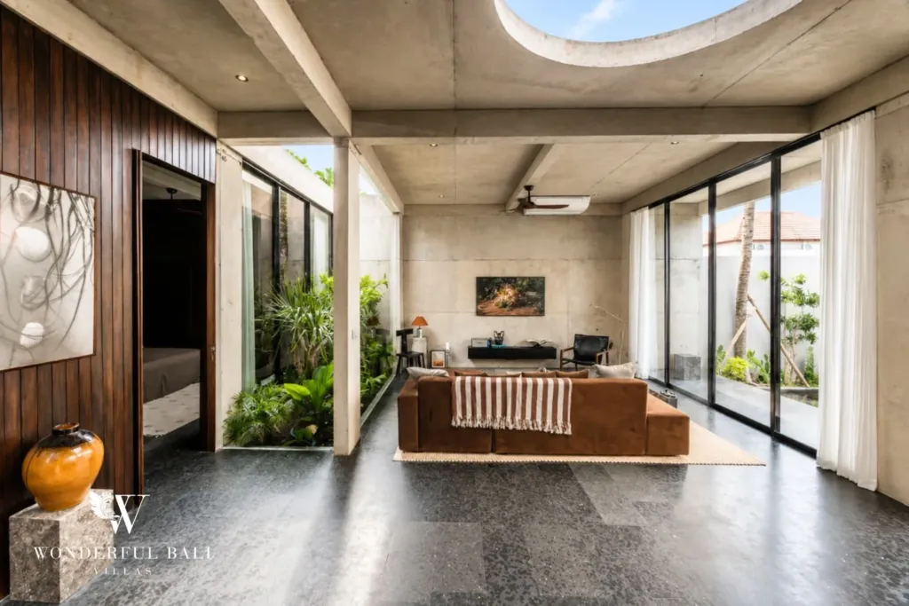 Living area with warm timber paneling, a brown sofa and an open doorway revealing a bedroom beyond, lit by full-height glass walls at a 2-bedroom villa in Seseh, Bali