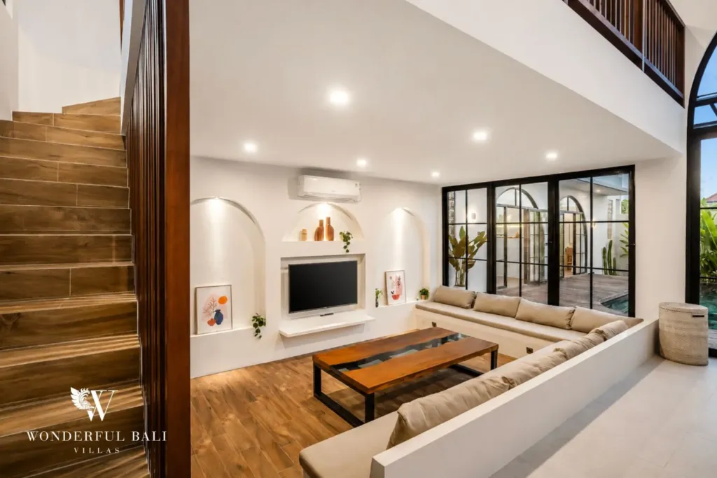 A cozy living lounge with beige sofas, arched wall niches, a wooden staircase, and steel-frame doors to the pool at a 3-bedroom villa in Pererenan, Bali