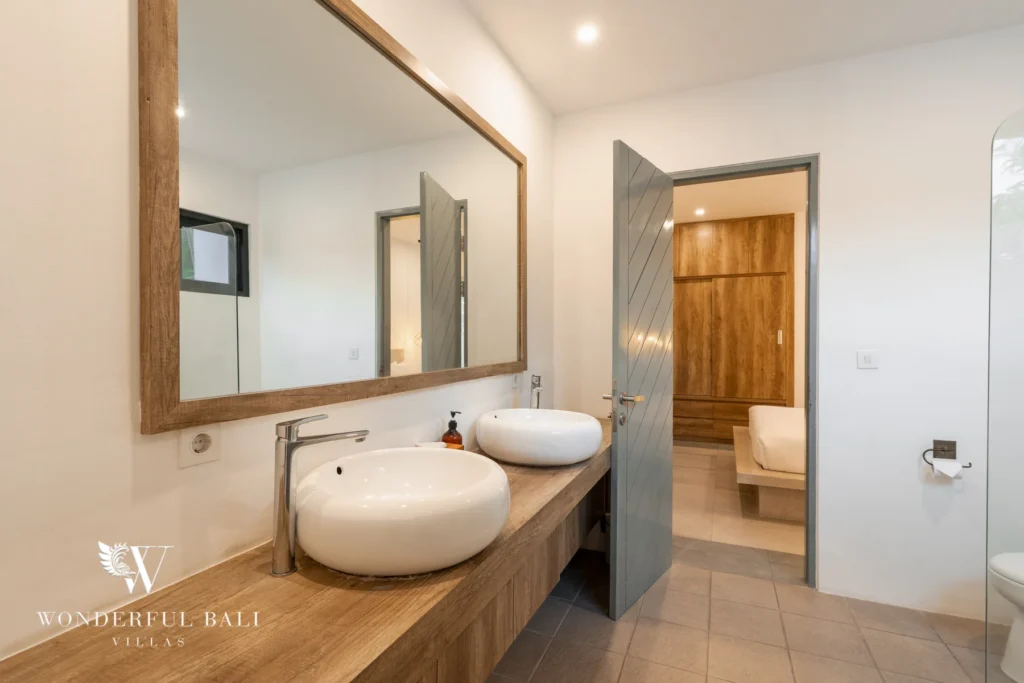 A bathroom with a wooden vanity holding twin vessel basins, a framed mirror and an open door revealing a wardrobe and bench at a 3-bedroom villa in Berawa, Bali