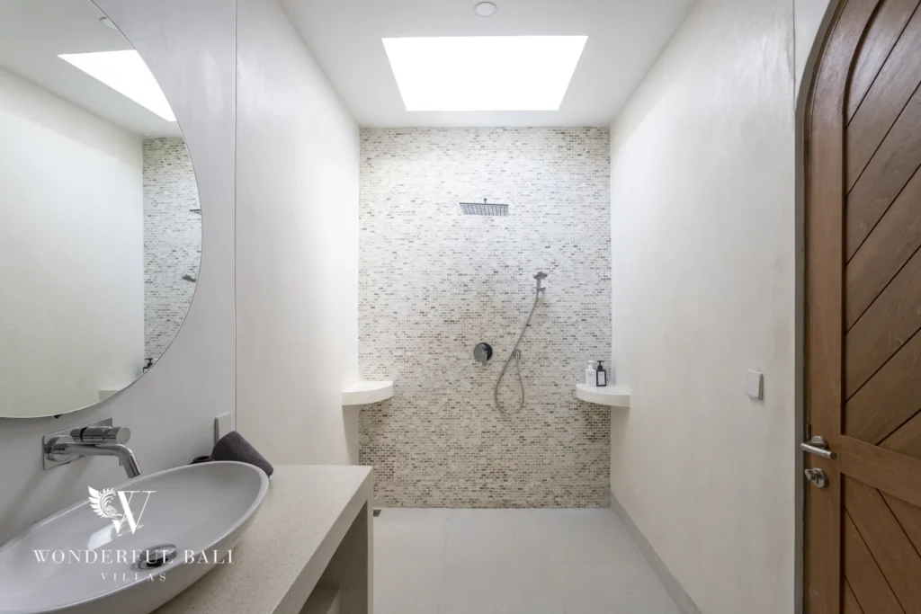 A white bathroom with a vessel sink, round mirror, skylight and mosaic-tiled walk-in shower beside an arched wooden door at a 3-bedroom villa in Bingin, Uluwatu, Bali
