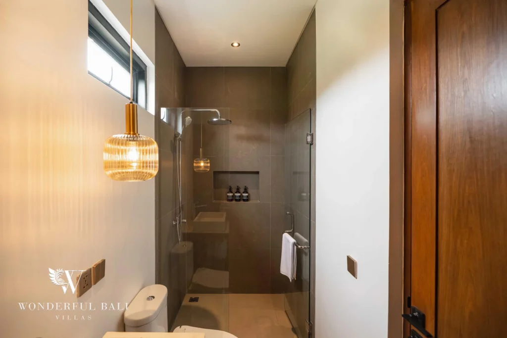 A narrow en-suite bathroom with grey-tiled walk-in glass shower, toilet, wooden door and a ribbed amber pendant lamp at a 4-bedroom villa in Canggu, Bali