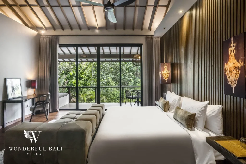 A bedroom with a white bed and velvet bench, exposed timber beam ceiling, a desk and floor-to-ceiling glass doors to a balcony at a 3-bedroom villa in Pererenan, Bali