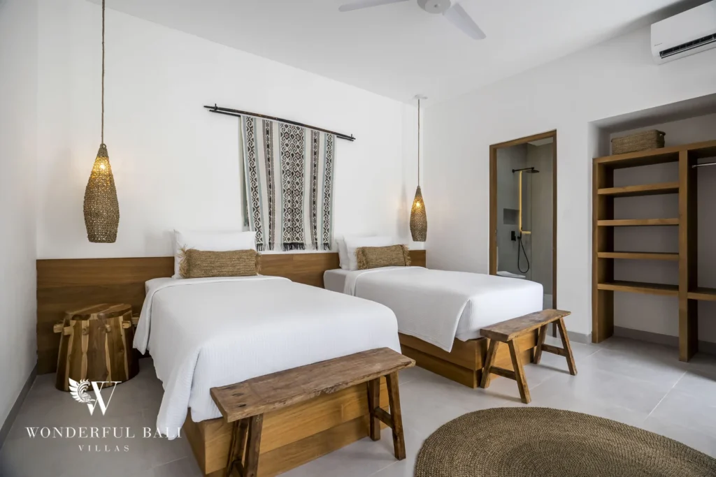 Two single beds with wooden benches and woven pendant lamps below a patterned textile hanging, with an en-suite shower and shelving alongside at a 3-bedroom villa in Bingin, Uluwatu, Bali