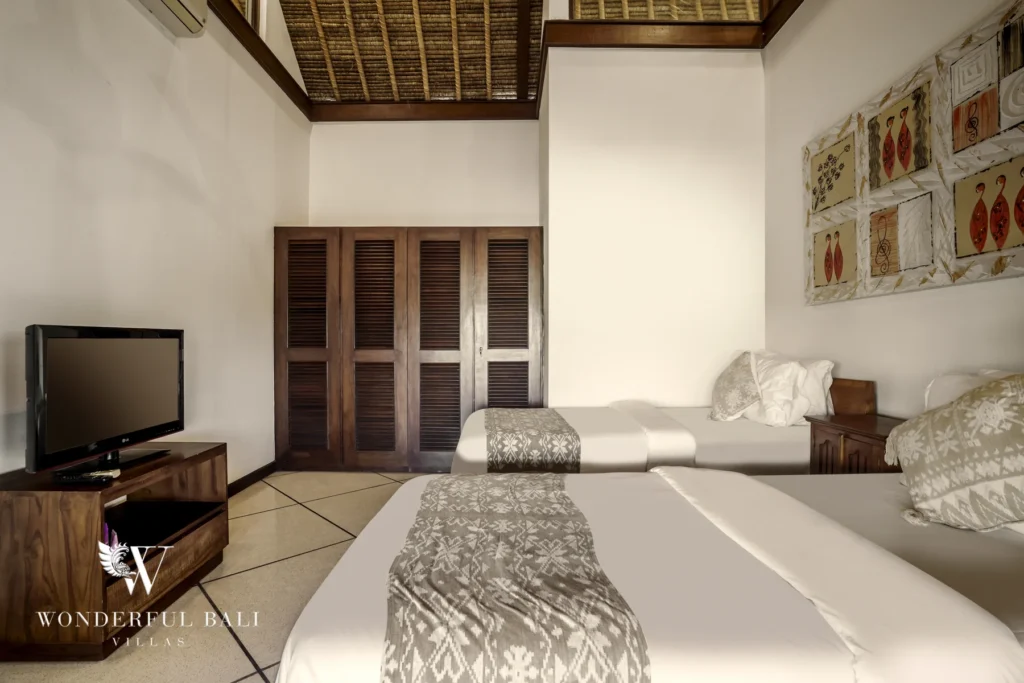 A twin-bed bedroom with a thatched ceiling, wooden wardrobe, television and framed wall art at a 4-bedroom villa in Seminyak, Bali