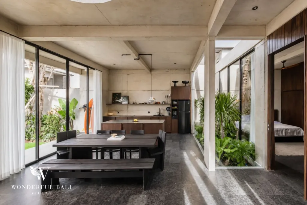Dining zone with a black timber table and bench beside the open kitchen, with a glazed courtyard and bedroom visible through the glass at a 2-bedroom villa in Seseh, Bali