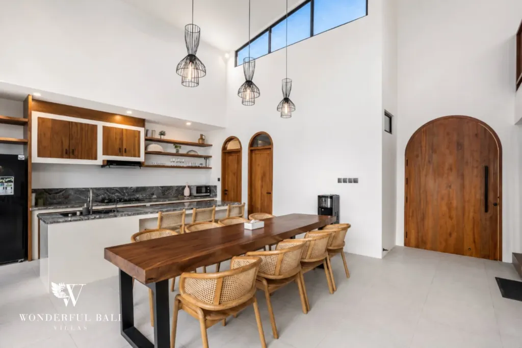 A live-edge wooden dining table with rattan chairs beside a wood-cabinet kitchen and an arched timber door at a 3-bedroom villa in Pererenan, Bali