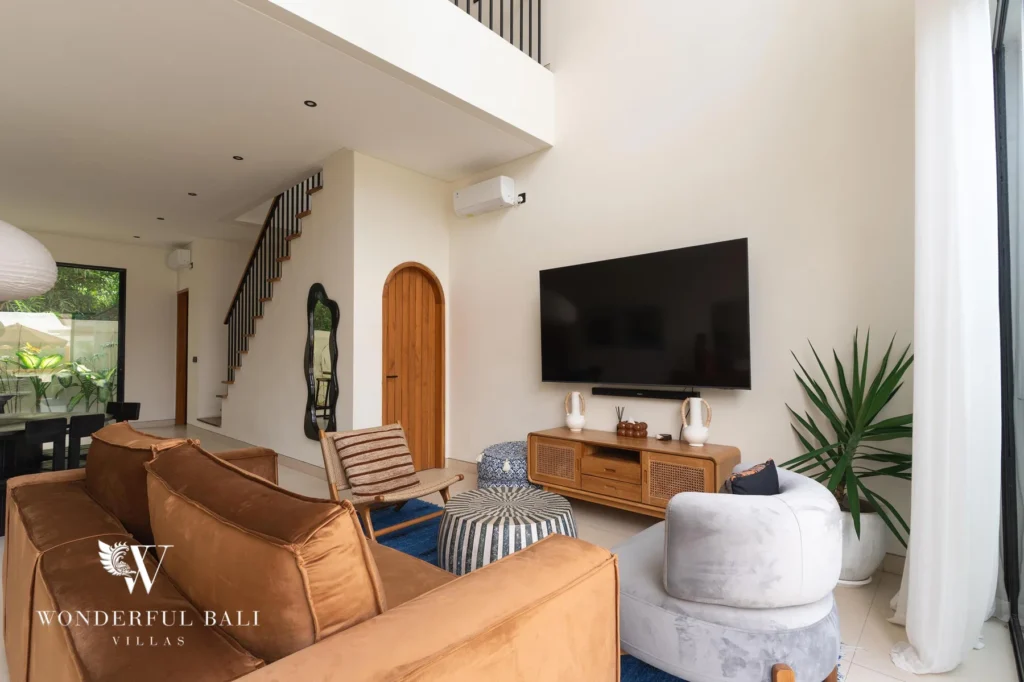 A double-height living room with a tan sofa, smart TV, staircase and arched door opening toward the garden at a 2-bedroom villa in Canggu, Bali