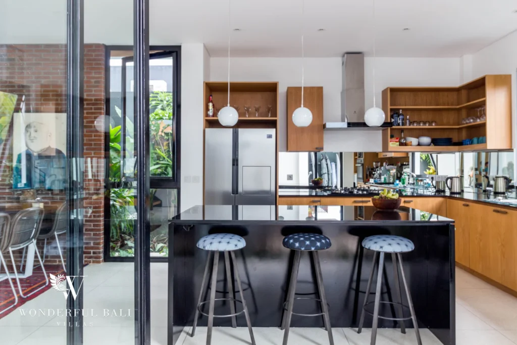 A kitchen with a black island and breakfast-bar stools, timber cabinetry and glass sliding doors onto a brick-walled garden at a 4-bedroom villa in Batu Bolong, Canggu, Bali