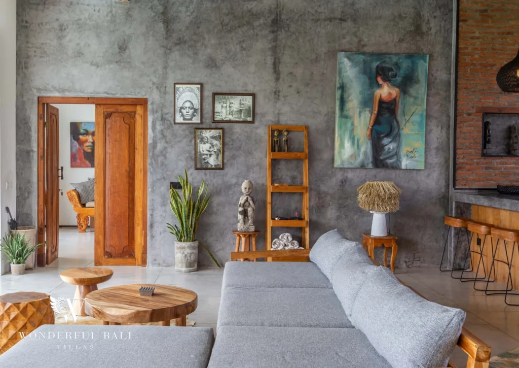 A concrete-walled living corner with grey sofas, Balinese art, a timber ladder shelf and a carved doorway leading to another room at a 3-bedroom villa in Seminyak, Bali