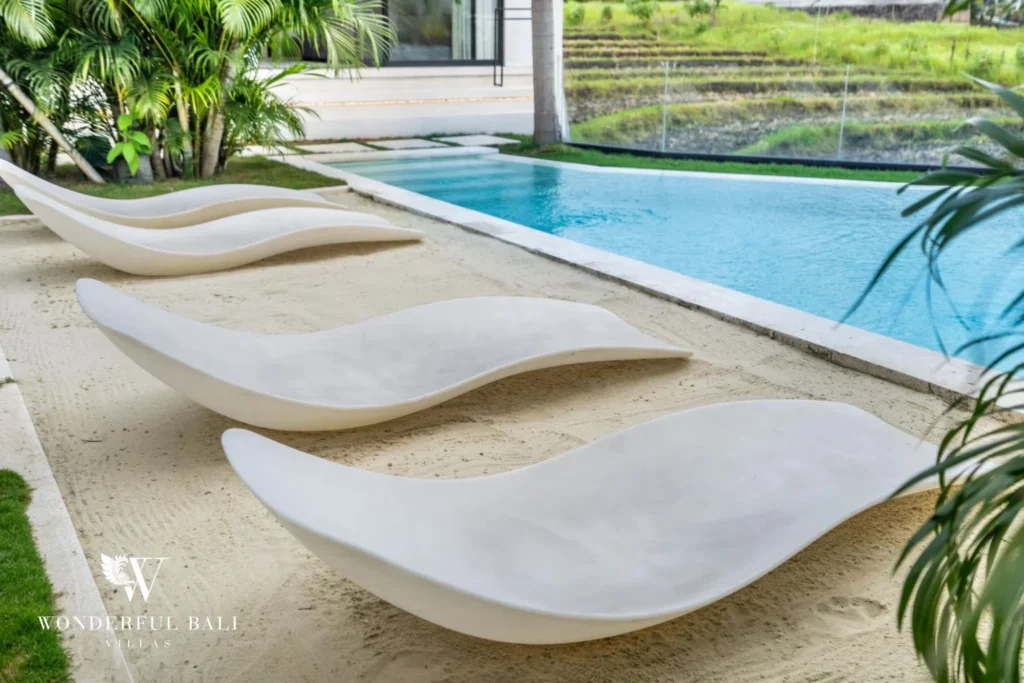 Sculpted white wave-shaped sun loungers set on sand beside the pool with rice-field views at a 5-bedroom villa in Pererenan, Bali