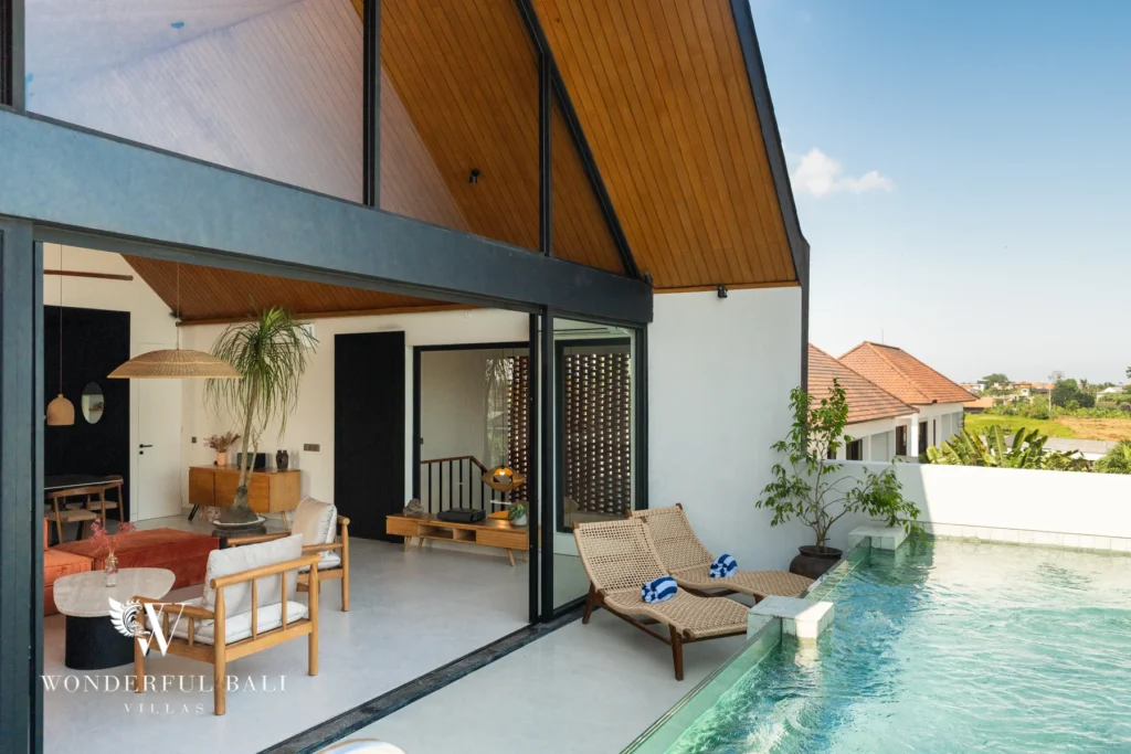 A covered outdoor terrace with timber loungers and armchairs beside a pool under a dramatic peaked wooden roof, with rice fields and rooftops beyond at a 5-bedroom villa in Batu Bolong, Canggu, Bali
