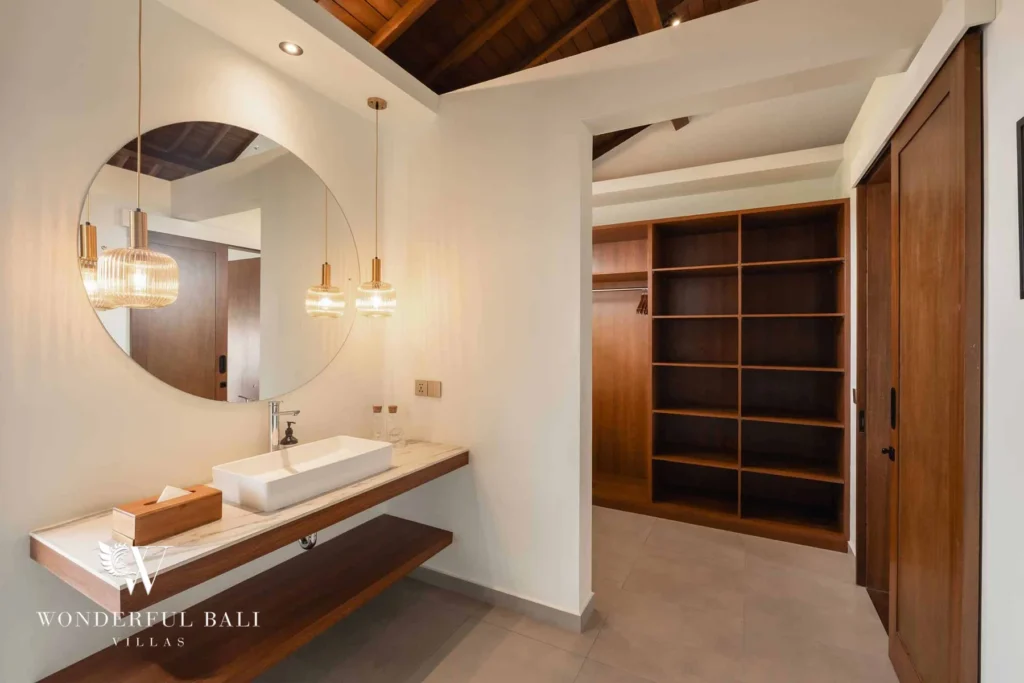 A bathroom vanity with a round mirror and pendants beside open wooden wardrobe shelving under a timber ceiling at a 2-bedroom villa in Canggu, Bali