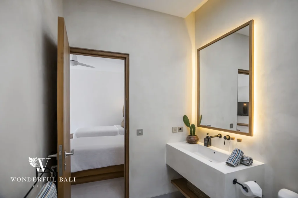 A minimalist bathroom with a floating stone sink, backlit mirror and a potted cactus, its door open onto a bedroom at a 3-bedroom villa in Bingin, Uluwatu, Bali
