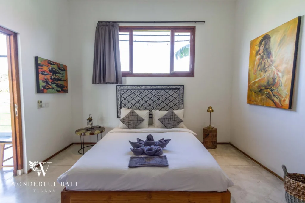 A bright bedroom with a carved headboard, white bedding folded into a flower, framed artworks and a curtained window at a 3-bedroom villa in Seminyak, Bali