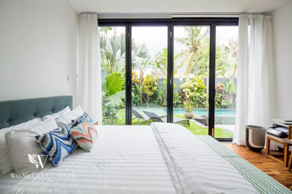 A bedroom with white bedding and sliding glass doors framing the pool and tropical garden beyond at a 4-bedroom villa in Batu Bolong, Canggu, Bali