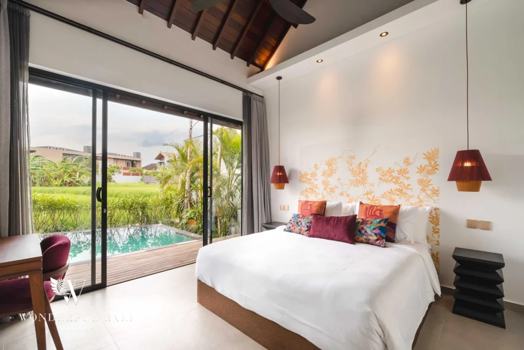 A bedroom under a vaulted timber ceiling with a floral gold headboard and red lamps, sliding glass doors opening to a private plunge pool and rice fields at a 4-bedroom villa in Canggu, Bali