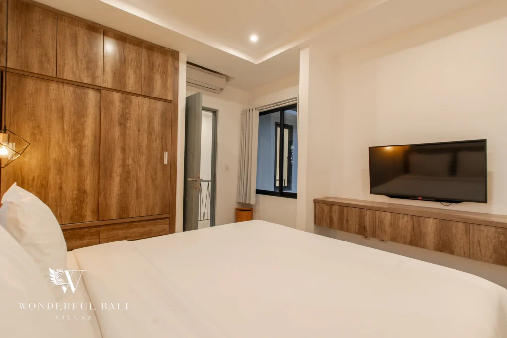 A bedroom with a double bed, tall wooden wardrobe, wall-mounted TV on a timber console and a window with grey curtains at a 3-bedroom villa in Berawa, Bali