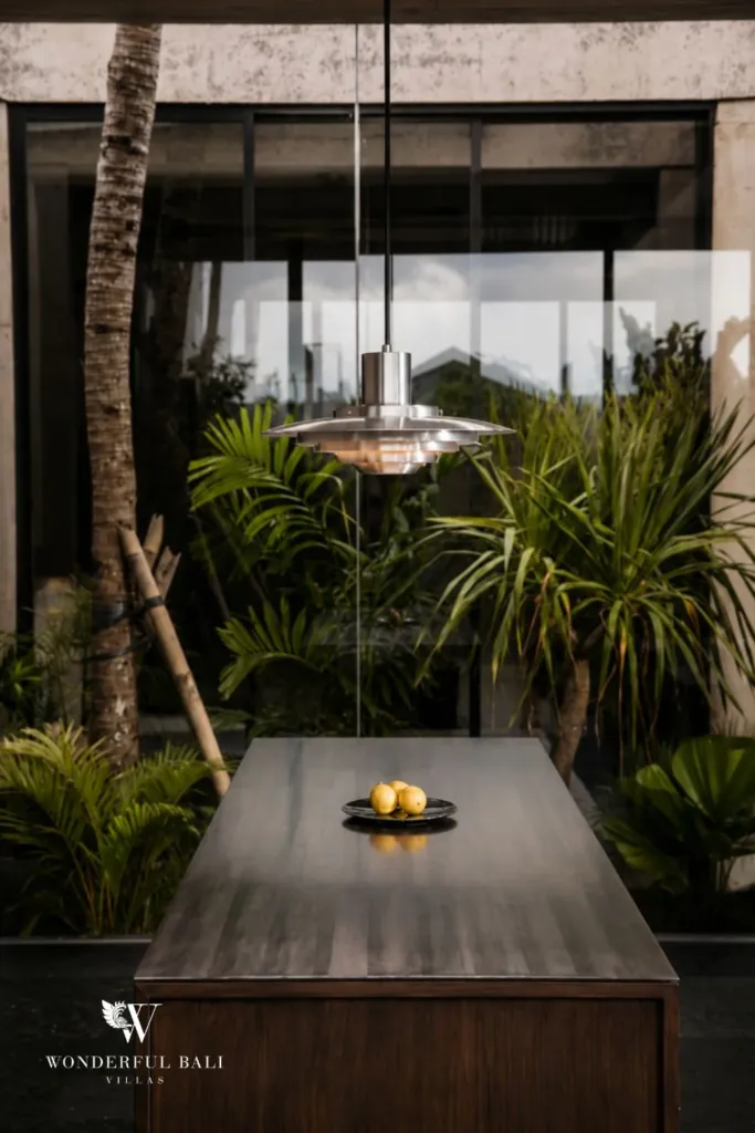Detail of the timber kitchen island topped with a bowl of lemons beneath a steel pendant lamp against a courtyard of palms at a 2-bedroom villa in Seseh, Bali