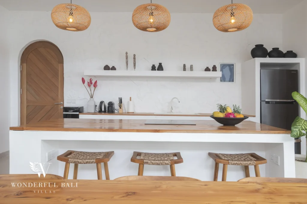 A kitchen island with a wooden countertop, woven bar stools, rattan pendant lights and an arched wooden door at a 3-bedroom villa in Bingin, Uluwatu, Bali