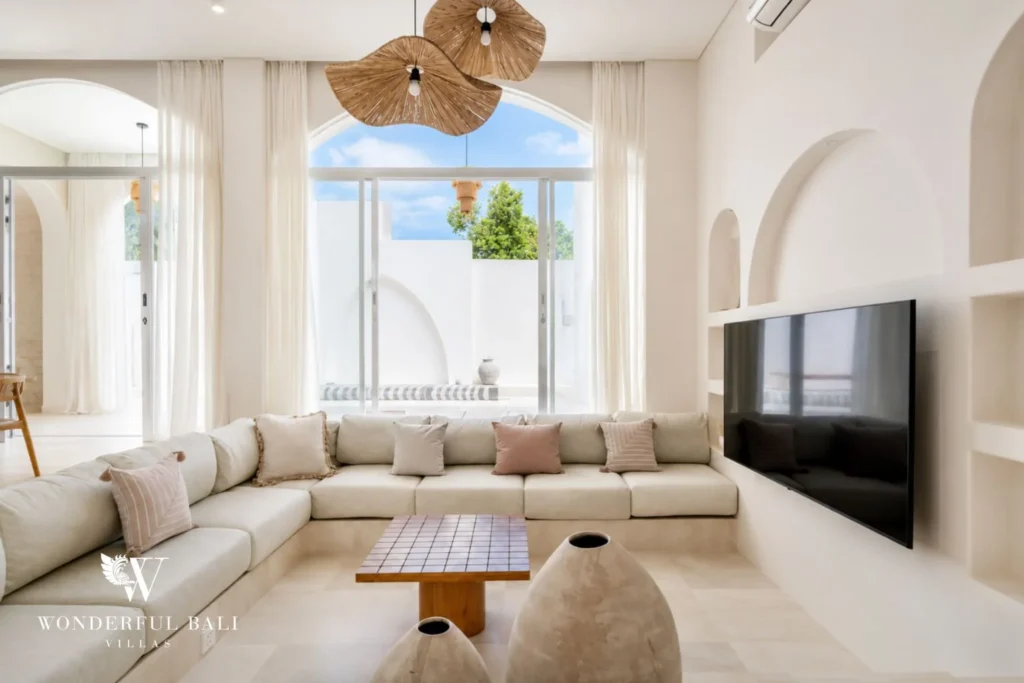 A sunlit living lounge with a low cream sofa, wall-mounted TV and a large arched window, at a 2-bedroom villa in Bingin, Uluwatu, Bali