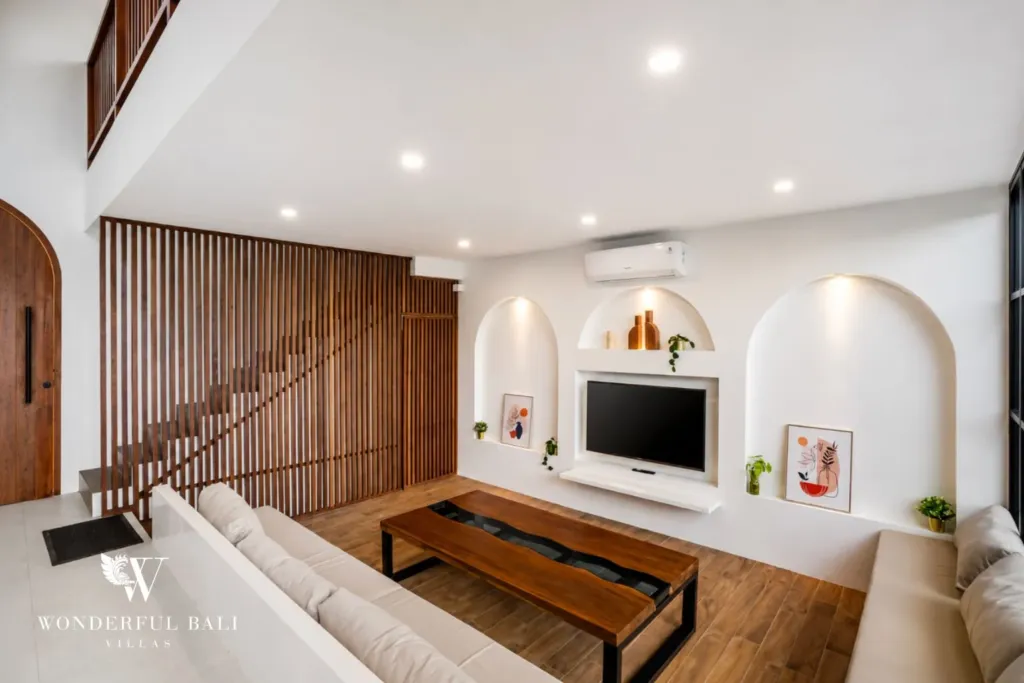 A white living room with arched display niches, wood-slat feature wall, and a staircase leading up to the mezzanine at a 3-bedroom villa in Pererenan, Bali