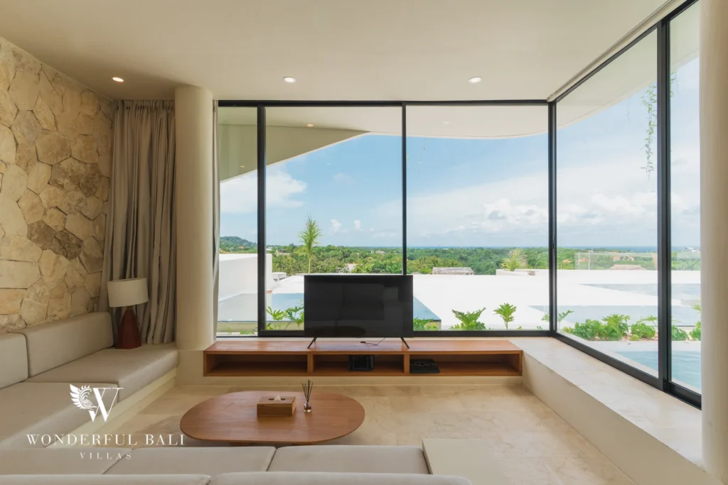 A living room with a stone feature wall, corner glass windows and a TV framing a panoramic view of greenery at a 3-bedroom villa in Bingin, Uluwatu, Bali