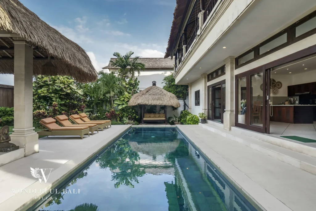 A blue swimming pool bordered by sun loungers, a thatched poolside cabana and the main building at a 4-bedroom villa in Seminyak, Bali
