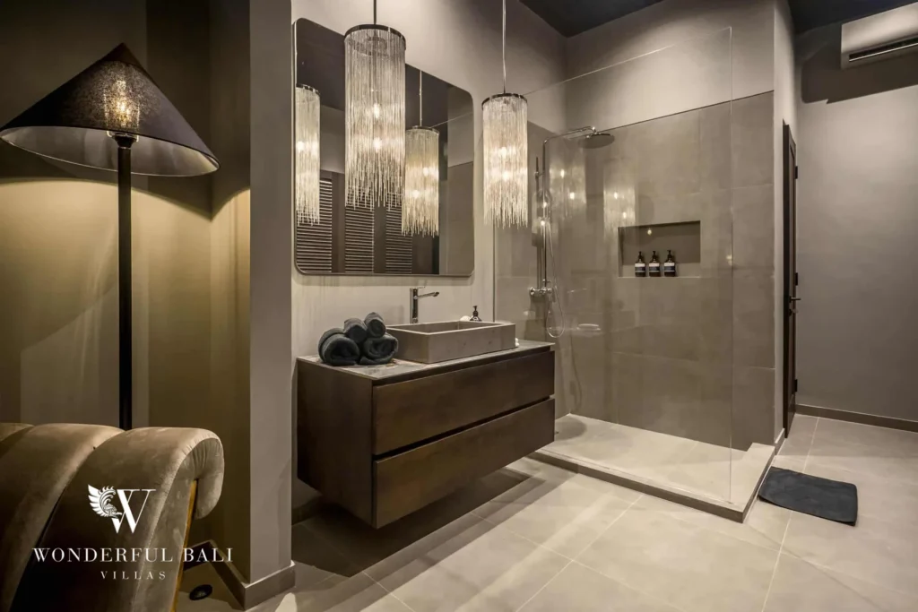 A grey-toned bathroom with a walk-in glass shower, a stone basin on a dark vanity, a floor lamp and layered pendant lighting at a 3-bedroom villa in Pererenan, Bali