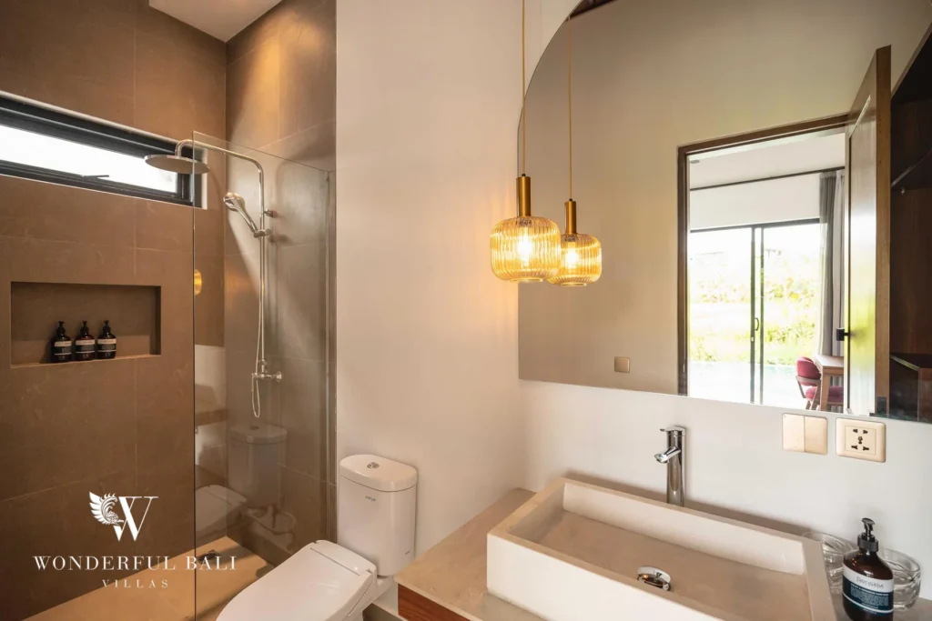 An en-suite bathroom with a stone vessel sink, arched mirror, twin amber pendant lamps and a grey-tiled glass shower at a 4-bedroom villa in Canggu, Bali