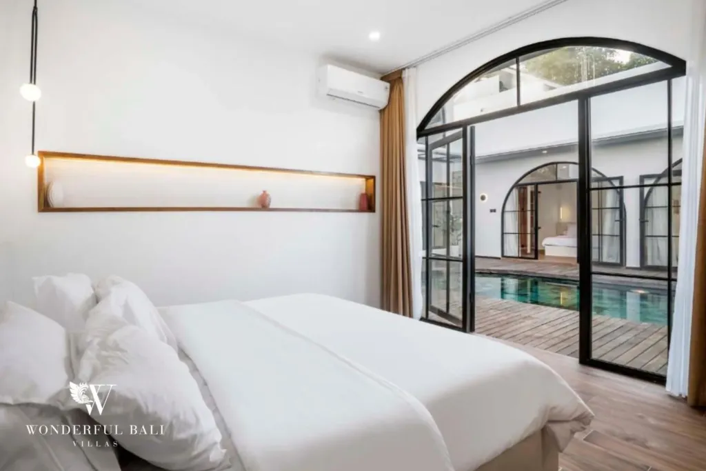 A bedroom with white bedding and a floating timber shelf opens through arched steel-frame doors to the pool at a 3-bedroom villa in Pererenan, Bali