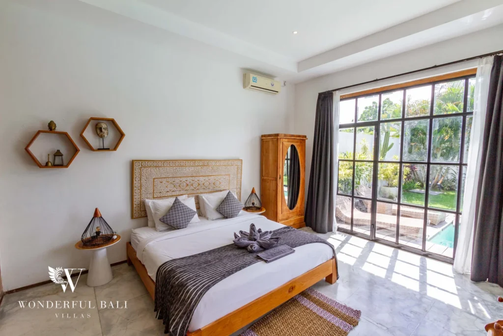 A bedroom with a carved wooden headboard, timber wardrobe and large steel-framed window overlooking the pool and garden at a 3-bedroom villa in Seminyak, Bali