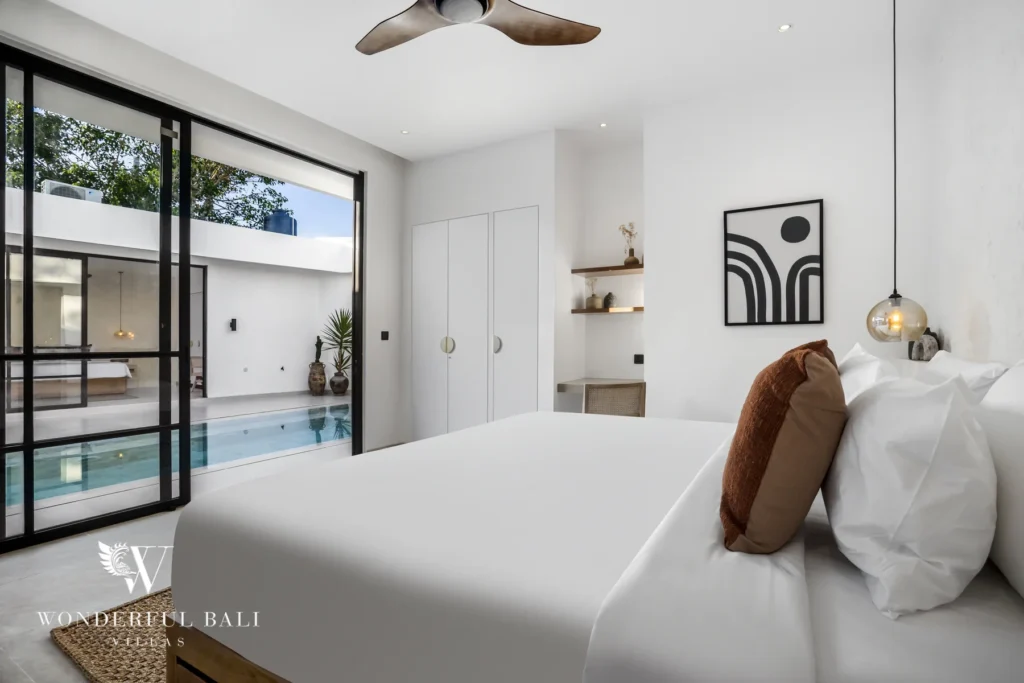 A bedroom with white bedding and a rust bolster cushion opening through glass doors to the poolside at a 3-bedroom villa in Bingin, Uluwatu, Bali