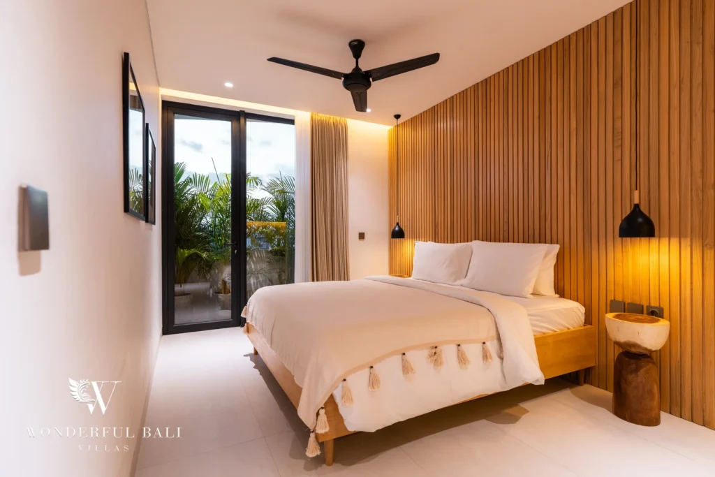 A serene bedroom with a slatted timber accent wall, white linens with tasseled throw, black pendant light and glass doors opening to greenery at a 5-bedroom villa in Batu Bolong, Canggu, Bali