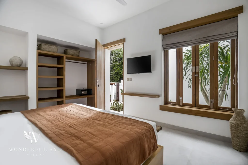 A bed with a rust quilt beside open wardrobe shelving, a wall-mounted TV and wood-framed windows and glass door to the garden at a 3-bedroom villa in Bingin, Uluwatu, Bali