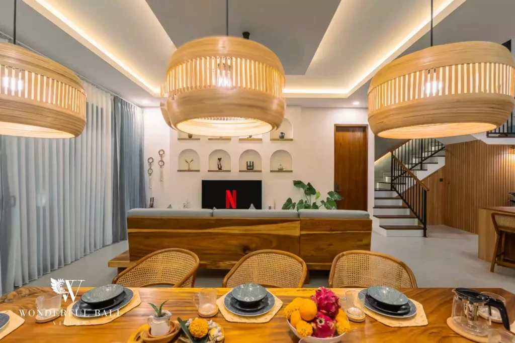 A timber dining table set for a meal under woven pendant lights with the living area behind at a 4-bedroom villa in Pererenan, Bali