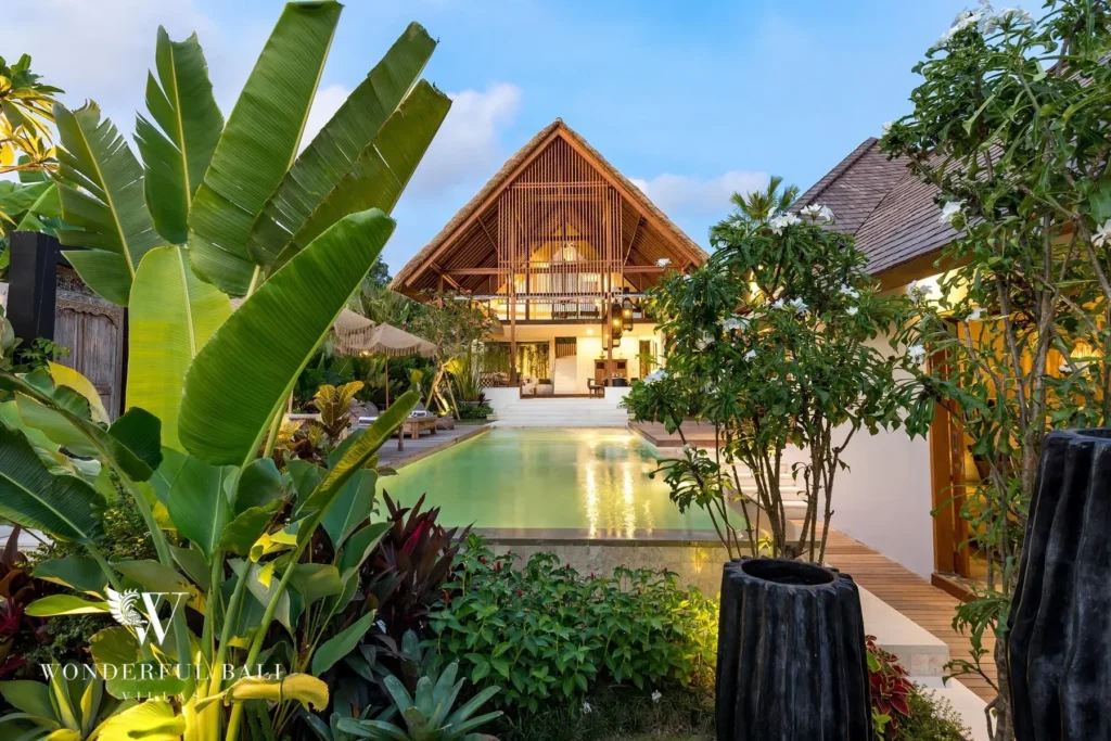 At dusk the lush garden frames the pool and the illuminated timber pavilion with its high pitched roof at a 4-bedroom villa in Canggu, Bali