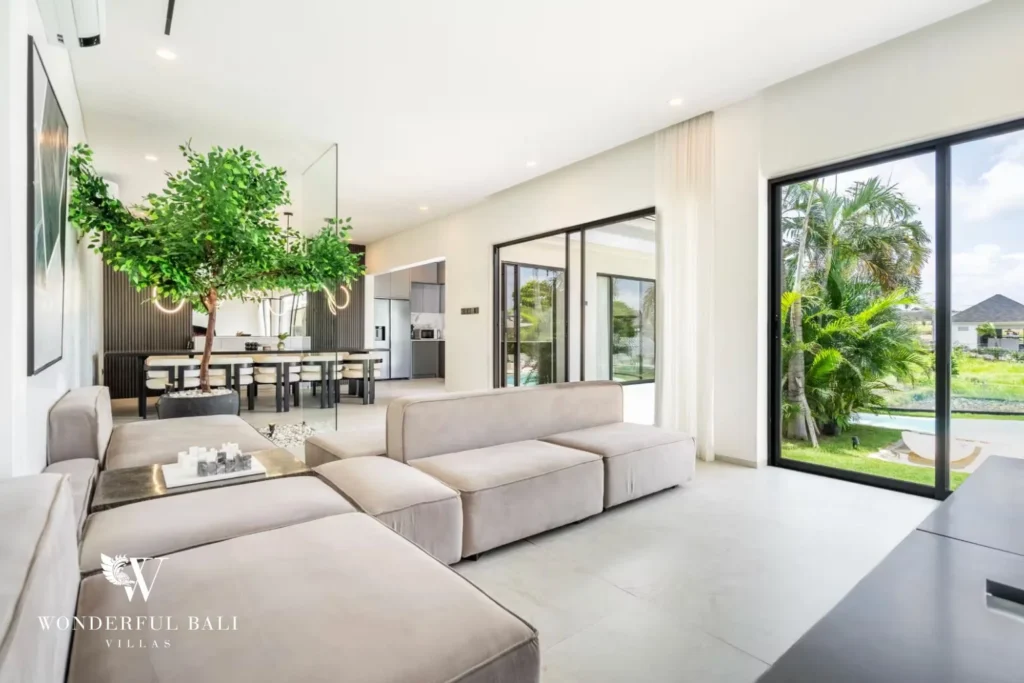 Open-plan living and dining space with a modular sofa, indoor tree and glass doors to the garden at a 5-bedroom villa in Pererenan, Bali