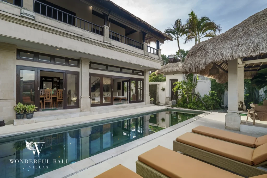 A pool running alongside the two-storey villa with open living areas and a thatched cabana under blue sky at a 4-bedroom villa in Seminyak, Bali