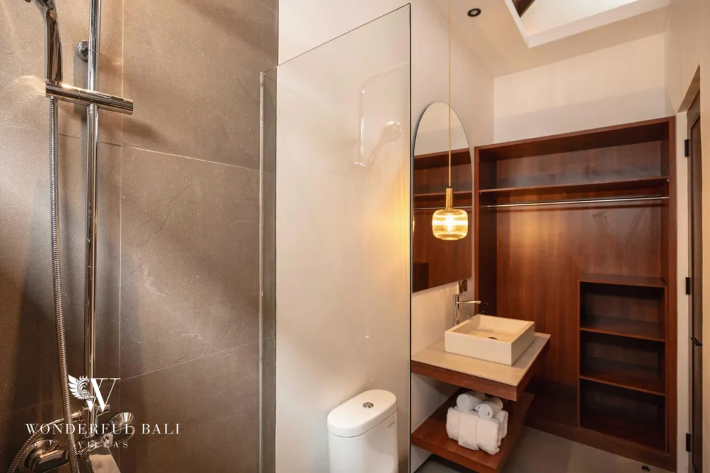 An en-suite bathroom with a grey-tiled glass shower, stone vessel sink and an adjoining timber open wardrobe with folded towels at a 4-bedroom villa in Canggu, Bali